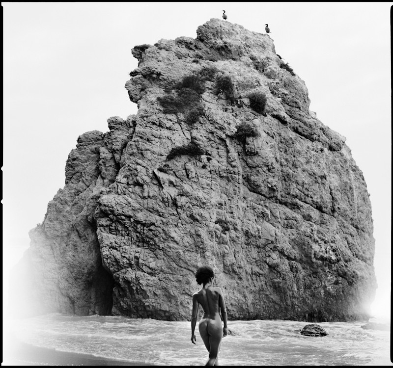 Nude woman on a beach walking towards  a large rock in California