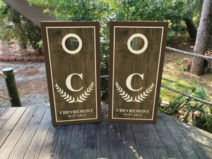 "Two cornhole board wraps with a rustic wood design, featuring a large initial 'C' surrounded by laurel branches and personalized text reading 'Chevremont' with a custom date, bordered with a clean frame, and white scoring holes near the top of each board." "Two cornhole board wraps with a rustic wood design, featuring a large initial 'C' surrounded by laurel branches and personalized text reading 'Chevremont' with a custom date, bordered with a clean frame, and white scoring holes near the top of each board."