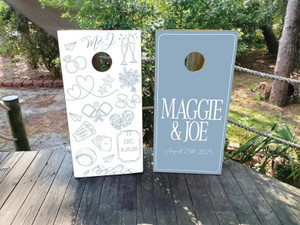 Dusty blue and white wedding cornhole board wraps featuring personalized designs for “Maggie & Joe – August 25th, 2025.” One board showcases elegant typography on a solid dusty blue background, while the other includes whimsical hand-drawn wedding illustrations such as rings, oysters, champagne flutes, hearts, and travel icons.