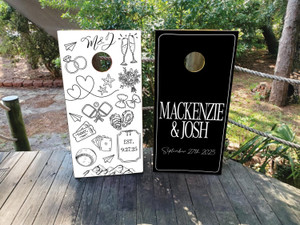 Modern black and white wedding cornhole board wraps featuring personalized couple names “Mackenzie & Josh” and date “September 27th, 2025.” One board showcases playful hand-drawn illustrations including rings, oysters, champagne glasses, and travel icons, while the other features a sleek minimalist design with elegant typography.