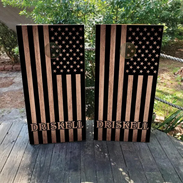 "Custom patriotic cornhole boards featuring a black and natural wood American flag design with a personalized monogram and last name. Perfect for military families, veterans, and patriotic celebrations."