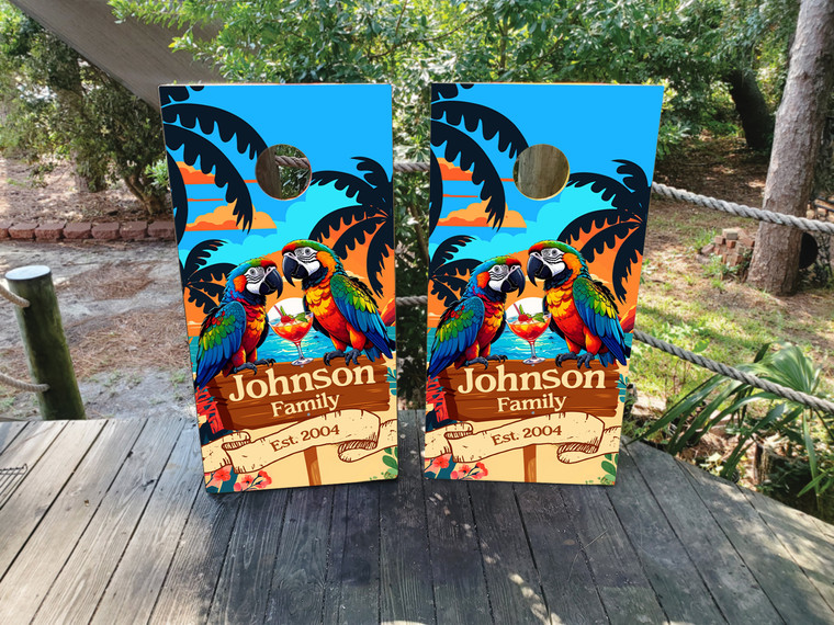 Colorful tropical-themed design featuring two vibrant parrots sitting beside a cocktail glass in front of a bright sunset with palm tree silhouettes, a blue sky, and personalized family name text on a wooden sign, printed as a full-board wrap design.