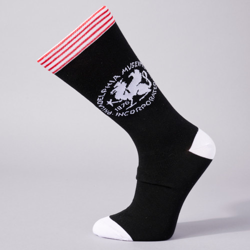Griffin socks are shown in side view on a sock form. The body of the sock is black, the toes and heels are white, and the top is striped in red and white. The griffin image in white is on the side of the sock and is circled with the words Philadelphia Museum of Art Incorporated, 1876.