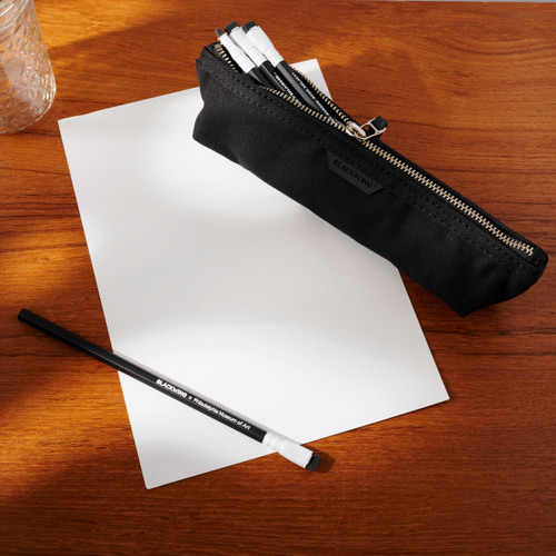 A half-zippered black canvas Blackwing pencil pouch sits diagonally across a sheet of white paper. A small number of black pencils with white ferrules and black erasers peek out from the top of the pouch. A single black and white Blackwing x Philadelphia Museum of Art pencil lies on the white sheet of paper. All of this sits on a wooden surface.