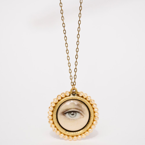 Detail of a round flat pendant featuring close up printed image of a single eye enclosed in resin and circled by tiny faux pearls around a gold-tone casing handing from a fine gold-tone chain