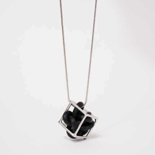 Detail of a pendant necklace featuring a hollow silver-toned cube encasing black rubber form suspended by smooth silver-toned chain