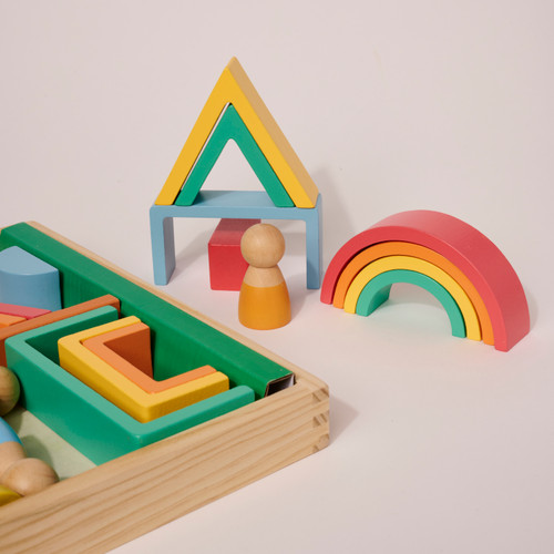 Zoomed in view of the corner of the block set crate with colorful blocks nested in it adjacent to a house and rainbow made of the wood blocks with a wood person standing next to them.
