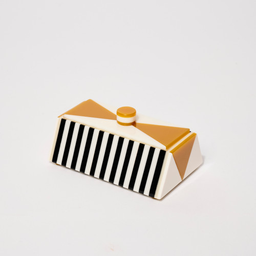 Rectangular acrylic box of vertical black and white stripes with triangular ends of ochre and cream and lift top of ochre and cream with small round knob of ochre and cream layers