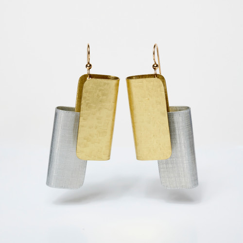 A pair of earrings made of a gold rectangular shape welded to a silver shape of the same style in an interlocking fashion, hanging in a white-box space.