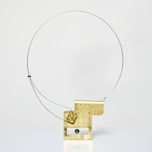 A gold collar-style necklace with a thin band and two gold rectangular shapes welded together asymmetrically, hanging in a white-box space.