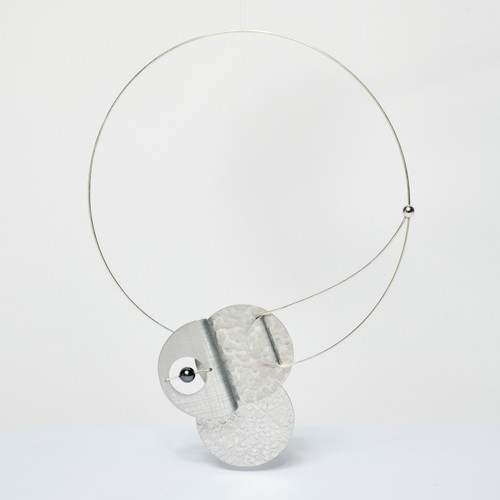 A thin, silver collar-style necklace with a large geometric overlapping closure at the front hanging in a white-box space.