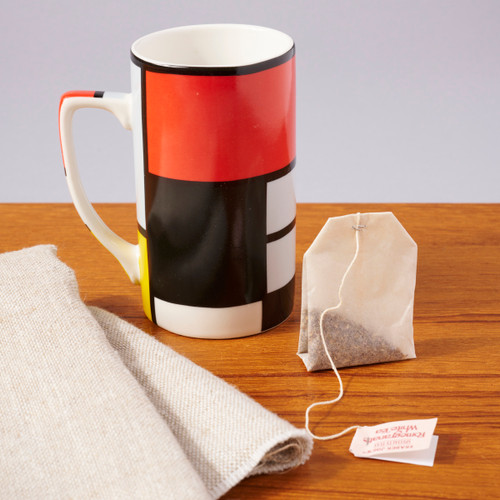 Image of a slender handled mug featuring red, black and yellow blocks of color and black lines of differing widths representing the art of Piet Mondrian. Sitting next to the mug is a tea bag and muslin tea towel.