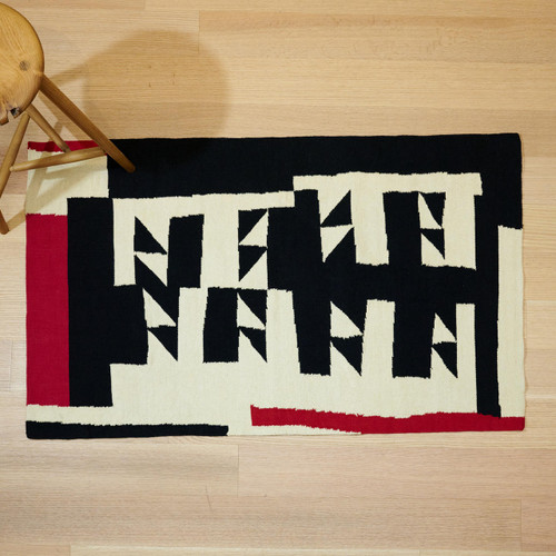 This rectangular cotton 3' x 5' rug is pictured in full view flat on a wooden floor,  against a wooden stool for scale. The rug is woven with a white background and a black angular design and  touches of red at one end and one side.