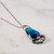 A blue teardrop shaped pendant is attached to an antique-finished gold filled chain. In this close-up view of the pendant, its teardrop center is shown in a bright blue beveled glass surrounded with antiqued metal. At the bottom of the pendant is a clear glass fan-shaped ornament.