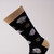 Arms and Armor helmet socks depict several styles of armor helmets in silver, interspersed with a beige motif. The body of the socks is black with beige toes and heels. One sock is shown in detail on a sock form in front and side view.