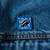 A pin of soft enamel in shades of blue over a base metal depicting a raven is shown pinned to a blue denim jacket.