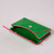 An emerald green envelope wallet with pink contrast edging lies flat and partially open on a grey surface. Its snap closure and interior are visible.