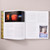 A spread of Lyle Ashton Harris: Our first and last love shows a photograph of a gallery with several illuminated photographs in shades of orange and blue above text on the left page and a photograph of a person sleeping in a bed under a black-and-white poster for the movie Caravaggio (1986).