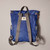 Back of Canfield Nylon Backpack in medium blue with grey handles and straps, and sewn on white manufacturer's logo at top near handle