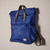 Side view of Canfield Nylon Backpack in medium blue with grey handles and straps, zip front patch pocket, and small white sewn on label on lower right of front showing depth