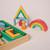 Zoomed in view of the corner of the block set crate with colorful blocks nested in it adjacent to a house and rainbow made of the wood blocks with a wood person standing next to them.