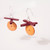 Side view of pair of coin-shaped orange disks with 2 dark red stick shaped beads suspended above with silver-toned ear wires