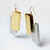 A pair of earrings made of a gold rectangular shape welded to a silver shape of the same style in an interlocking fashion, hanging in a white-box space at a 45-degree angle.