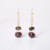 Front view of a pair of dangle earrings made of a gold shaft leading to an earthy gem encircled with red crystals.