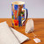 The Klee Burggarten mug sits atop a wooden surface in side view, next to a linen cloth and an open teabag. The mug's exterior is multicolored, in blocks of color. The handle is multicolored with a beige interior, the interior of the mug is beige.