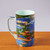 Monet Pond with Waterlilies mug is shown on a wooden surface in full view, handle to the left. The mug's exterior is in deep blues and greens showing lilies on a pond in the impressionist style, the handle also in blues and greens. The interior of the mug is pale green.