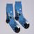 Side view of Rousseau Carnival Socks lying flat. The socks depicts bare trees in black against a nighttime dark blue sky with white stars. Toes and heels are black. Toward the toe stand two figures in white carnival dress.
