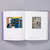 Interior spread of American Modernism: Highlights from the Philadelphia Museum of Art. The left page shows a colorful painting of two figures at a desk in a library. The right page shows a black and white woodcut image of a man operating a jackhammer with city buildings in the background.