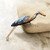 A pin of embroidery thread and beads in the shape of a blue heron is shown leaning against a stone on a marbled tabletop.