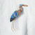 A pin of embroidery thread and beads in the shape of a blue heron is shown pinned to a piece of white woven fabric.