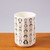 Sixty-five artists' images and names in black cover a simple white cup. The cup is handleless and sits stop a wooden surface.