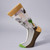 Side view of Sheeler Cactus socks on a sock form, depicting a cactus plant in a pot with its spikes removed,  sitting on a pedestal and surrounded by lights, ready for  photography. The socks have green toes,  are beige at the top; the image is in greens and browns.  The front and side of the socks are visible, showing brown soles and beige heels.