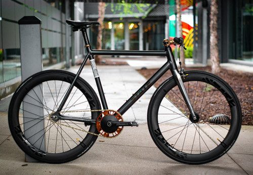 Minimalist black fixed-gear bicycle with orange crank and sleek black tires on urban sidewalk, perfect for commuting with the OpenPath Payments bike-friendly system.