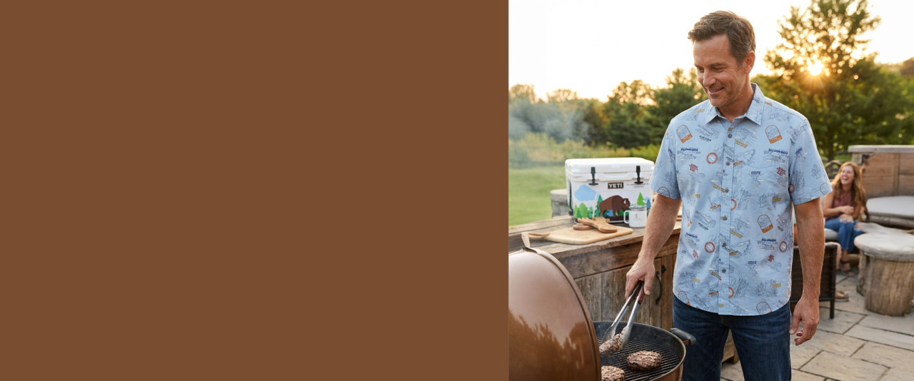 Man grilling hamburgers wearing a blue National Parks shirt with a yeti cooler, salad tongs, and woman in the background.