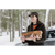 A woman carrying logs outside in a wooded area. She is wearing a brown plaid Button Up Cap, an orange shirt and an olive jacket.