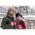 Photo of man and woman standing with arms around each other, holding coffee mugs in front of a large porch with stone railing and lots of windows in the winter. Man is wearing Stormy Kromer Insulated Waxed Cotton Cap in tan with olive wool coat. Woman is wearing red and black check jacket and black cloche style hat.