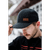 Photo of a man standing in front of a cabin, looking down and holding the brim of his dark gray plaid Curveball Cap. He is also wearing a black hoodie.