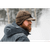 Side profile view of a man with a red beard wearing a dark brown waxed Rancher Cap with the earband pulled down over his ears. It is a very snowy scene with trees and blowing snow.