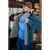 Photo of man putting on his green plaid Camp Shirt Jack over a bright blue shirt with a trout image. He is wearing a brown Stormy Kromer cap and is standing in front of a wood pile.