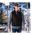Man wearing a cap and wool vest over light gray shirt walking in town.