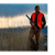 Man wearing a blaze orange vest, holding a shotgun, in a tall grass field.