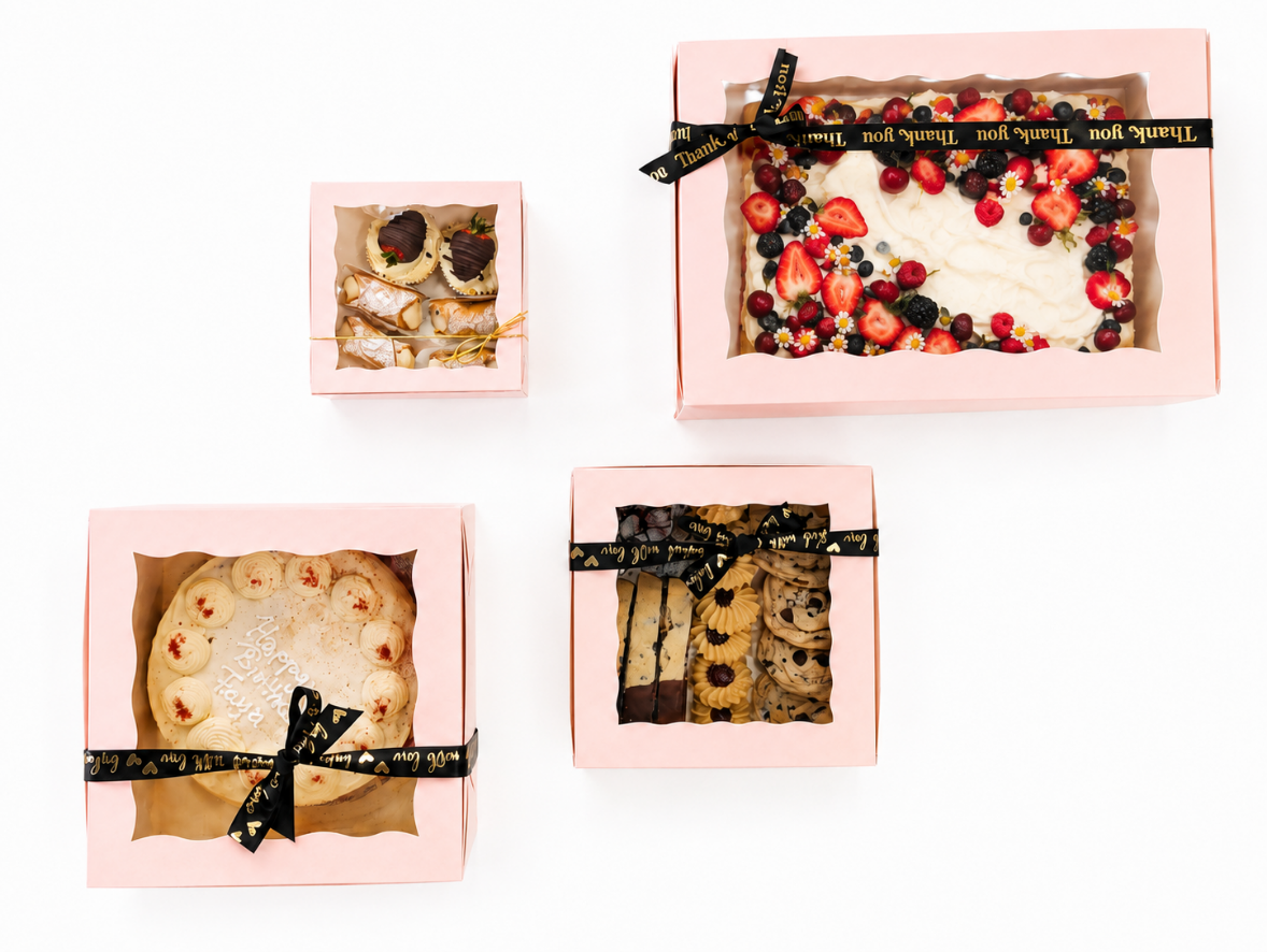 A collection of soft pink window bakery boxes filled with cakes, cookies, pastries, and decorated desserts displayed on a clean white background.