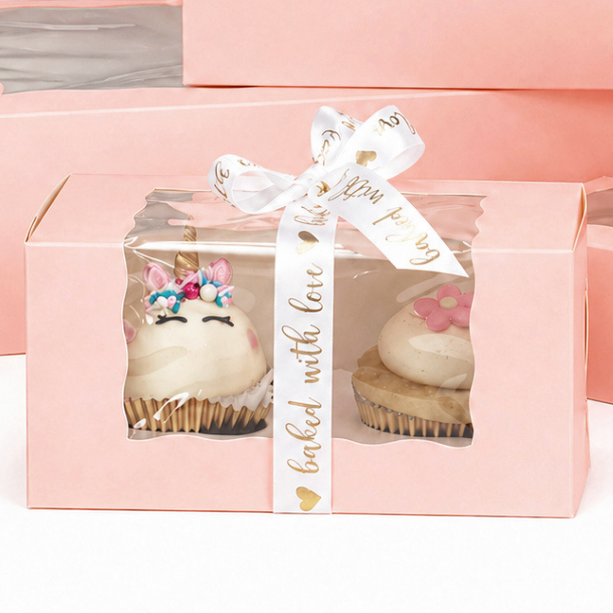 A pink window cupcake box holding two decorated cupcakes with ribbon, styled as a neat small-order bakery gift box.