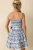 The Prosperina Miss Darcy Dusty Blue Floral Cotton Mini Dress is a charming statement piece designed with feminine details throughout. Crafted from crisp cotton fabric, this fit-and-flare mini dress features a dusty blue and ivory floral print, a smocked bodice for a comfortable and flattering fit, functional side pockets, and a square neckline framed by wide straps with scalloped trim accents. A coordinating self-tie waist sash defines the silhouette, while the tiered skirt creates playful movement with every step. Finished with scalloped trim detailing throughout, this polished mini is perfect for showers, garden parties, vacations, and sunny-day celebrations.