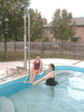 Aquatic Access IGAT-180 pool lift in use, assisting a woman into the pool while another person supports her