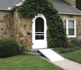 Solid curb ramp leading to a white door of a stone house, surrounded by greenery